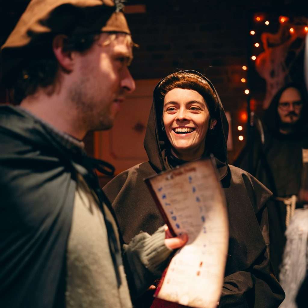 CC Clocktower--13 - Chance & Counters Two people in hooded medieval robes smile and chat, a parchment scroll visible in the foreground; warm lights in the background.