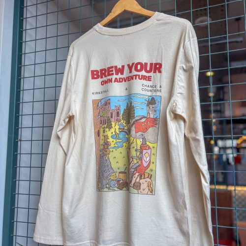 IMG_2458 - Chance & Counters Beige long-sleeve shirt on a wooden hanger with a colorful graphic and the words 'BREW YOUR OWN ADVENTURE' displayed on the front.