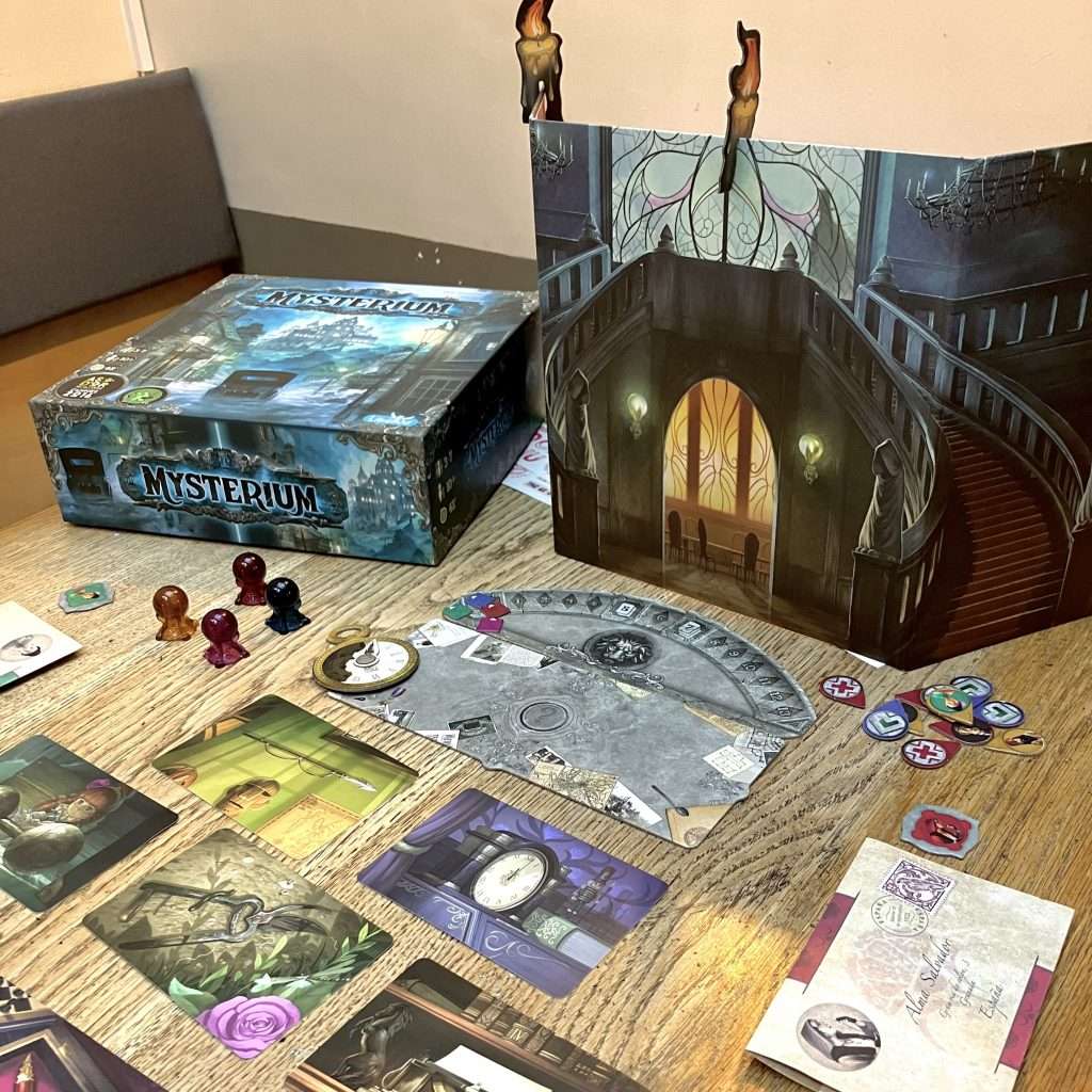 IMG_7150 - Chance & Counters Tabletop board game setup featuring the Mystarium box, a fold-out dark stairwell board, and scattered cards and token discs.