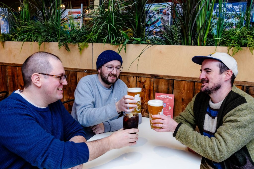PHOTO-2026-05-14-18-57-31 - Chance & Counters Three friends raise beers in a toast at a white table, smiling in a casual indoor setting with wooden paneling and plants in the background.