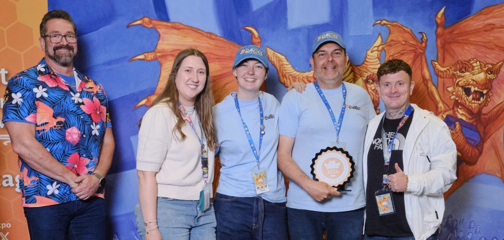 Patrick Campbell award - Chance & Counters Five people pose for a group photo at an event, two in light blue shirts with lanyards, one holds a round award plaque, backdrop shows a blue dragon mural.