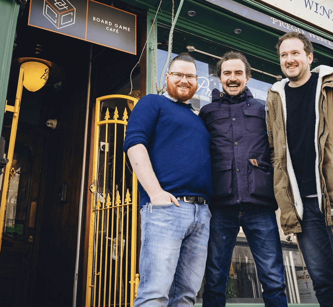Screenshot 2026-05-14 at 141923 - Chance & Counters Three men pose together outside a green storefront with a Board Game Cafe sign and Italian Wine Bar above them, smiling at the camera.