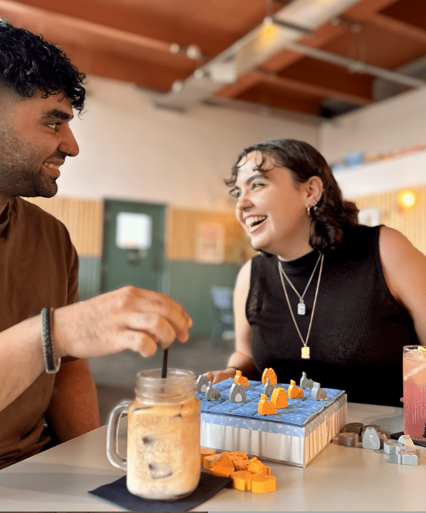 Two friends smiling and chatting while playing a board game at a table, with a blue game box and orange/gray pieces in front of them.