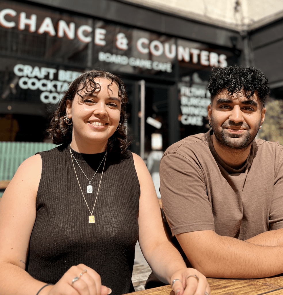 Two friends sit at a wooden table outside a board game cafe, smiling at the camera.