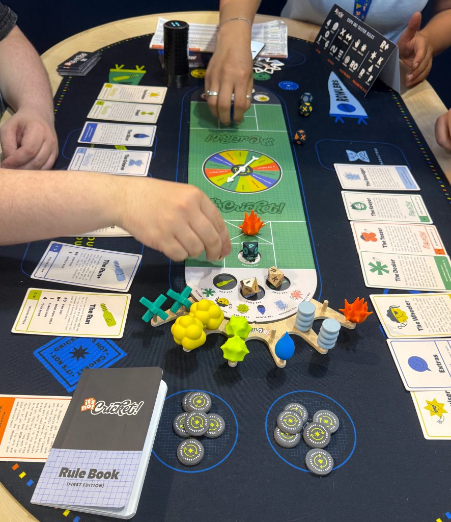 Tournament 3 - Chance & Counters People playing a cricket-themed board game with a green game mat, cards on the sides, dice, and player pieces arranged around a spinner wheel at the center of the table.
