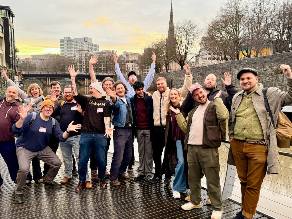 WhatsApp Image 2026-01-21 at 105538 1 - Chance & Counters A diverse group of people on a wooden boardwalk by the river, smiling and raising their hands at the camera with city buildings in the background.