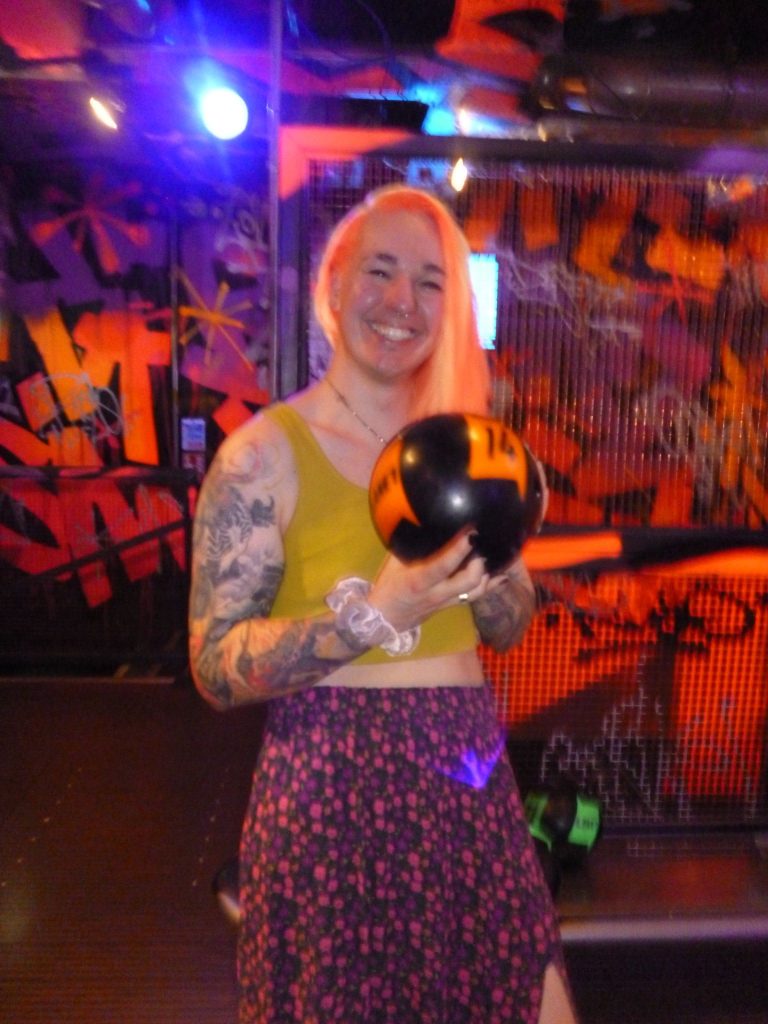 Smiling person with pink hair wearing a yellow top, holding a black-and-yellow helmet in a neon-lit graffiti-filled venue.