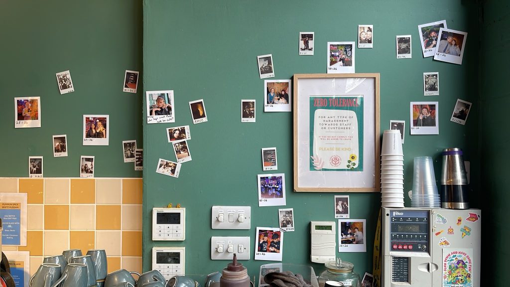 Wall of Polaroid photos taped to a green wall with a framed 'Zero Tolerance' sign near a coffee station. (informative)