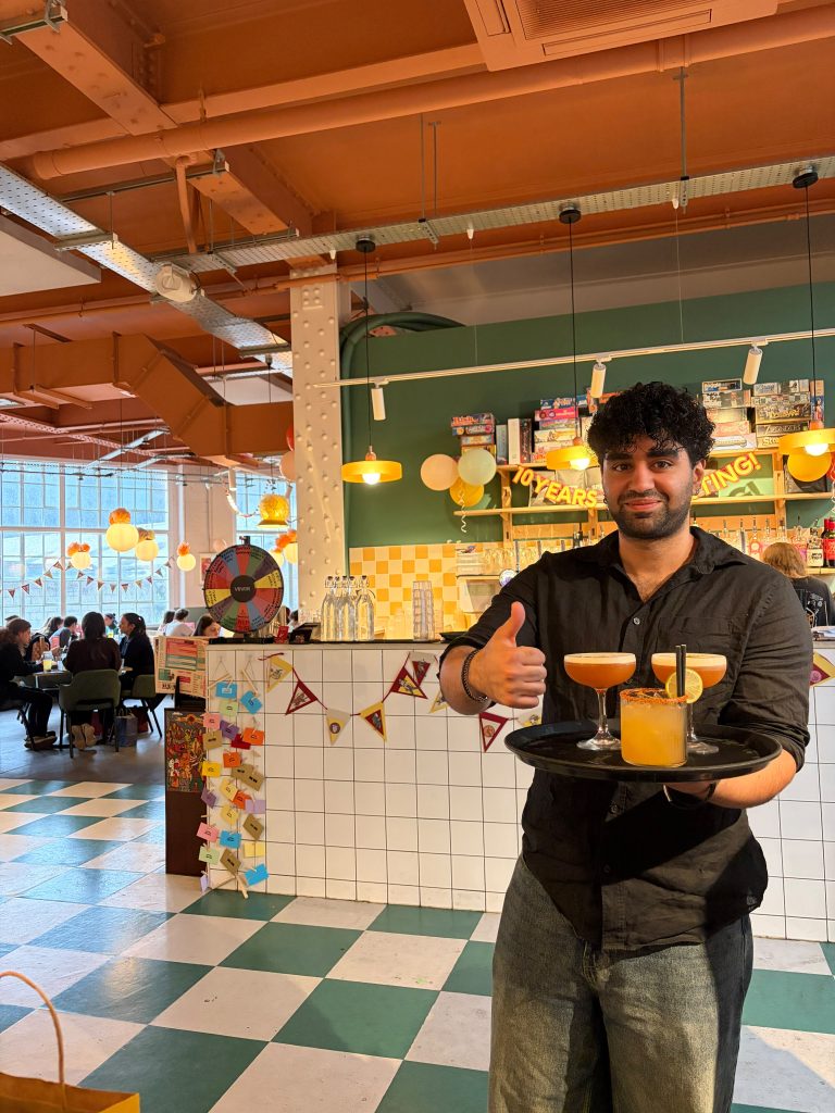 Smiling server giving a thumbs up while holding a tray with two cocktails and a juice, in a bright cafe setting.