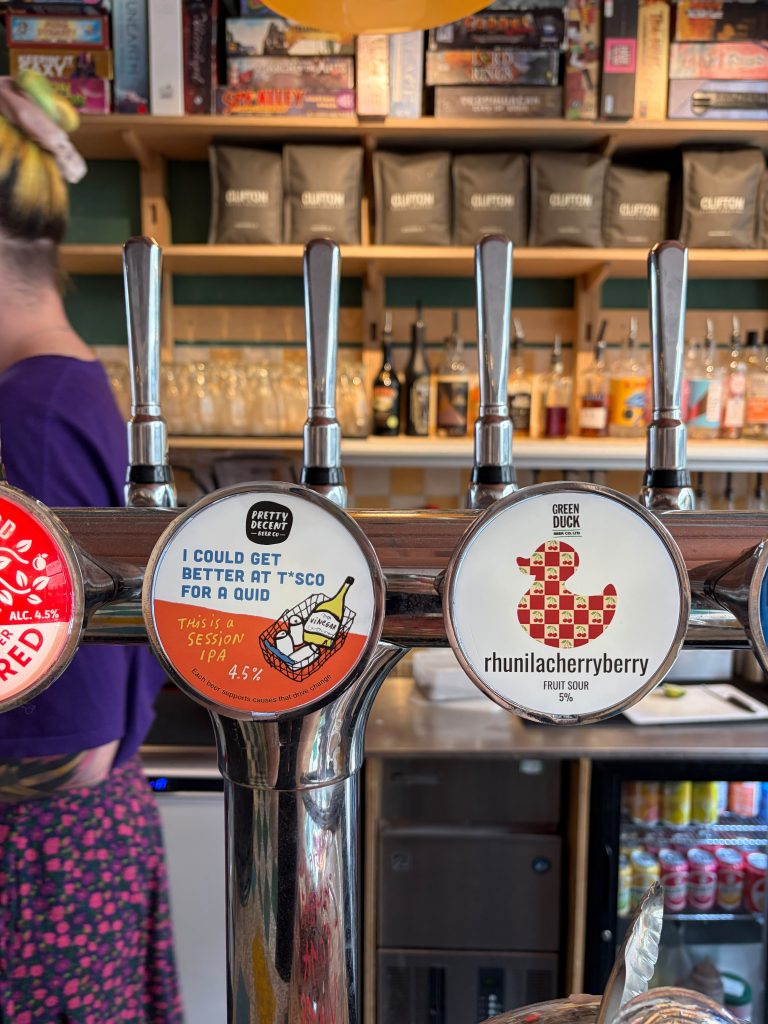 Row of beer taps with circular label designs in a bar, a person in purple on the left and shelves in the background.