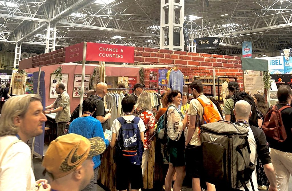Busy convention floor with a brick-walled booth labeled 'Chance & Counters' and people browsing clothes and merchandise in racks.