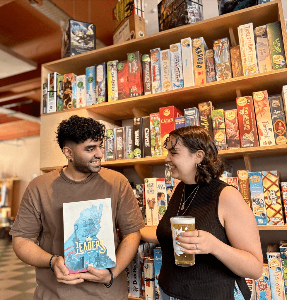 Two people smile at each other in a board game store, shelves stacked with colorful game boxes behind them; the man holds a graphic novel titled'Leaders'.